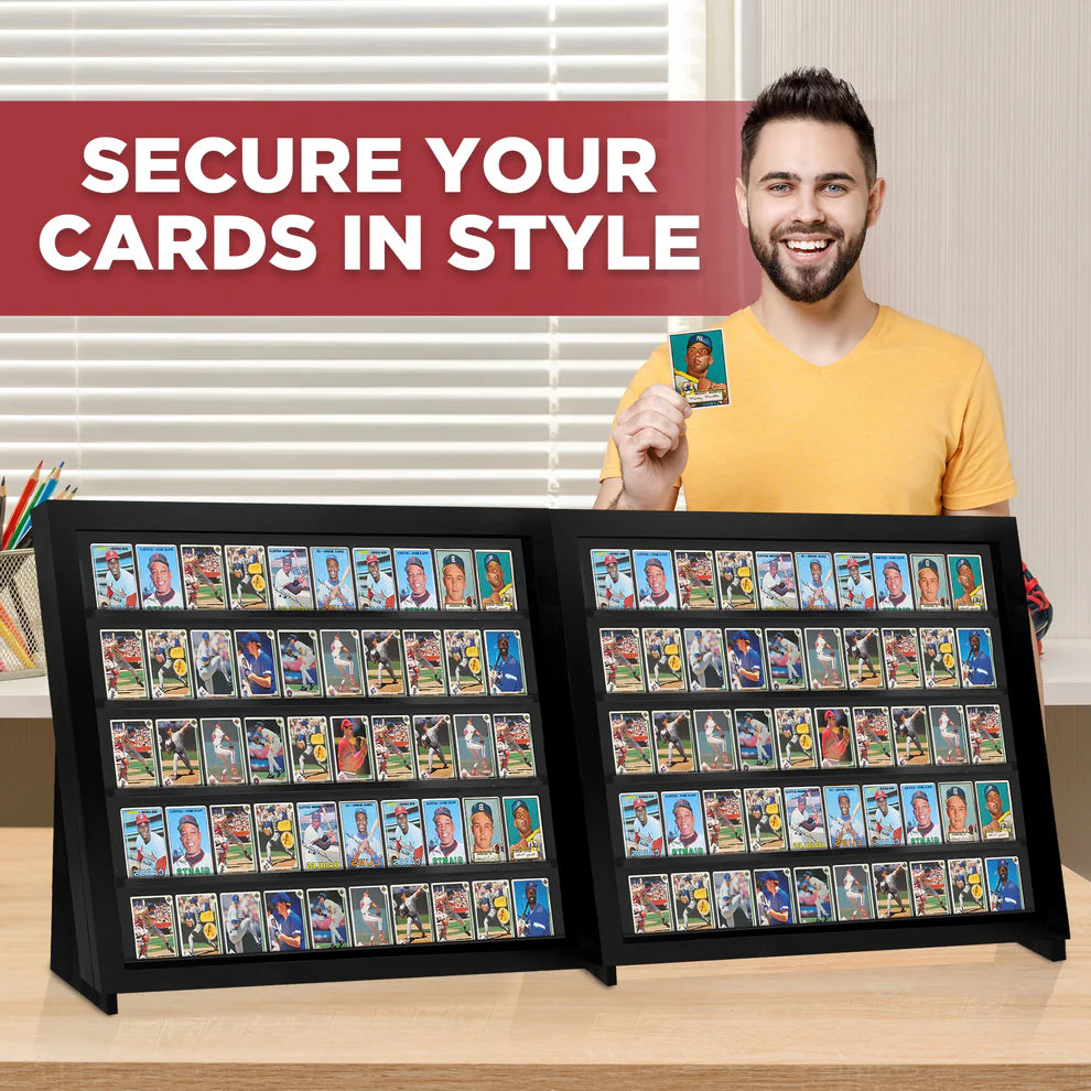 Man holding a trading card with two large display cases filled with cards on a wooden surface.
